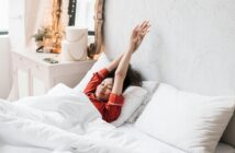 Frau in rotem Pyjama, die im Bett liegt und die Arme in die Höhe streckt.