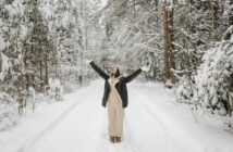 Frau steht auf winterlichem Weg im Wald und reckt die Arme in die Höhe.